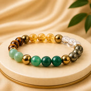 A beaded stretch bracelet displayed on a round beige pedestal. The bracelet features a mix of translucent amber-yellow beads, metallic gold beads, clear quartz, brown tiger's eye, light green jade, dark green malachite, and metallic silver beads.