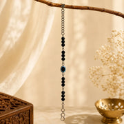 A long beaded hanging charm with black round beads, a central blue evil eye motif, and silver-toned chain ends, displayed against a warm cream backdrop with soft shadows and decorative props.